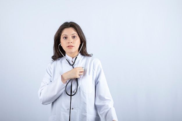 Young female doctor using stethoscope to check pulse on white wall. 