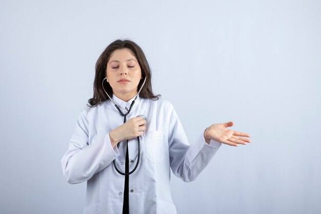 Young female doctor using stethoscope to check pulse on white wall.