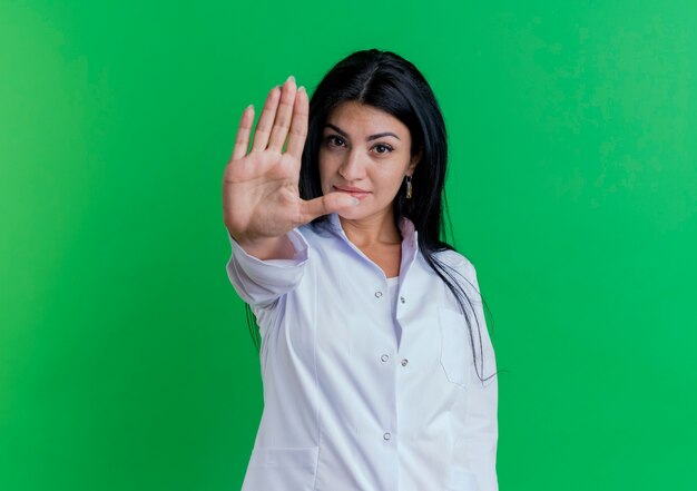 Young female doctor wearing medical robe  gesturing stop isolated on green wall with copy space