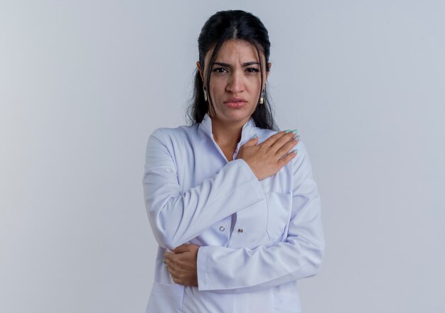 Young female doctor wearing medical robe looking putting hand on shoulder suffering from pain isolated