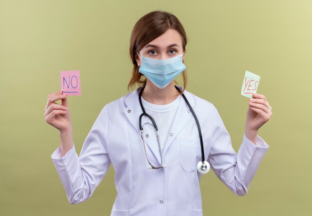 Young female doctor wearing medical robe, mask and stethoscope holding yes and no notes and looking  on isolated green wall