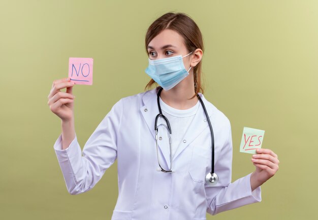 young female doctor wearing medical robe, mask and stethoscope holding yes and no notes and looking at no note on isolated green wall