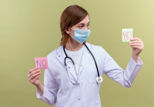 Young female doctor wearing medical robe, mask and stethoscope holding yes and no notes and looking at yes note on isolated green wall