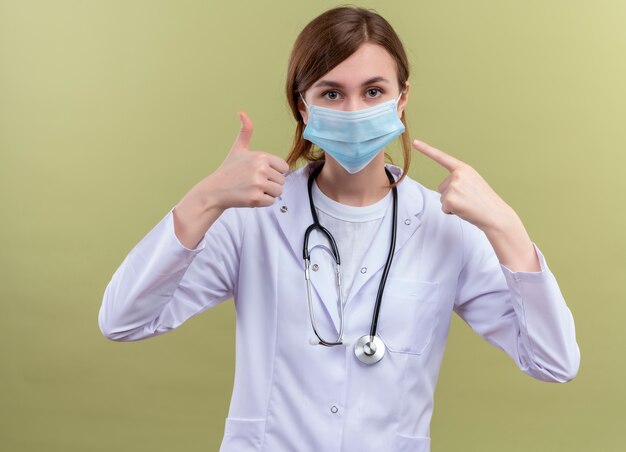 Young female doctor wearing medical robe, mask and stethoscope showing thumb up and pointing at her mask on isolated green wall