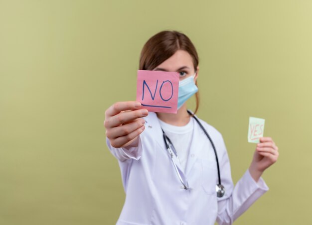 young female doctor wearing medical robe, mask and stethoscope stretching no note  on isolated green wall with copy space