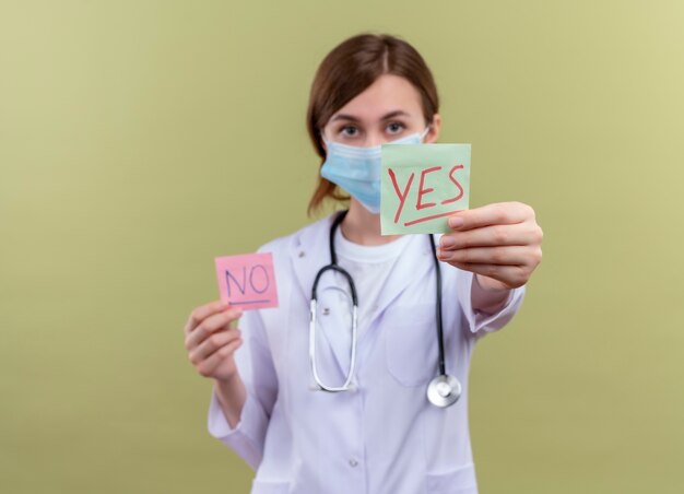 Young female doctor wearing medical robe, mask and stethoscope stretching yes note  on isolated green wall with copy space