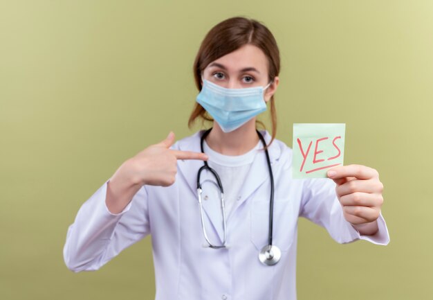 Young female doctor wearing medical robe, mask and stethoscope stretching yes note and pointing at herself on isolated green wall