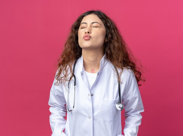 Молодая женщина-врач в медицинском халате и стетоскопе делает жест поцелуя с закрытыми глазами, изолированными на розовой стене
