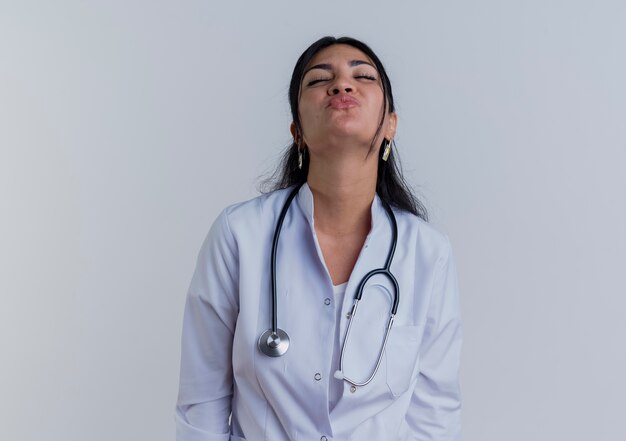 Young female doctor wearing medical robe and stethoscope doing kiss gesture with closed eyes isolated