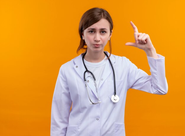 コピースペースと孤立したオレンジ色の壁にサイズジェスチャーを行う医療ローブと聴診器を身に着けている若い女性医師