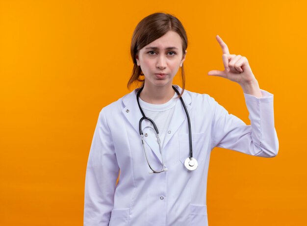 Young female doctor wearing medical robe and stethoscope doing size gesture on isolated orange wall with copy space