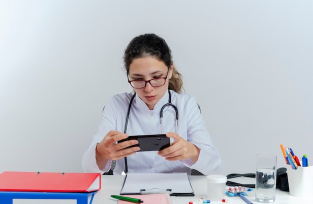 Young female doctor wearing medical robe and stethoscope and glasses sitting at desk with medical tools using mobile phone isolated