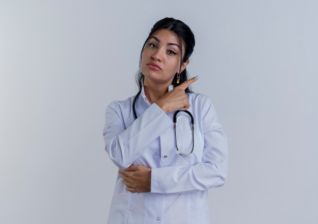 Young female doctor wearing medical robe and stethoscope looking pointing at side isolated