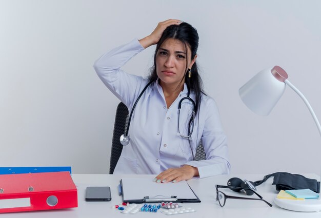 Молодая женщина-врач в медицинском халате и стетоскопе сидит за столом с медицинскими инструментами и смотрит, кладет руки на стол и на голову с изолированной головной болью
