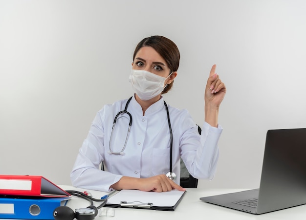 Young female doctor wearing medical robe with stethoscope in medical mask sitting at desk work on computer with medical tools points to side on white wall with copy space