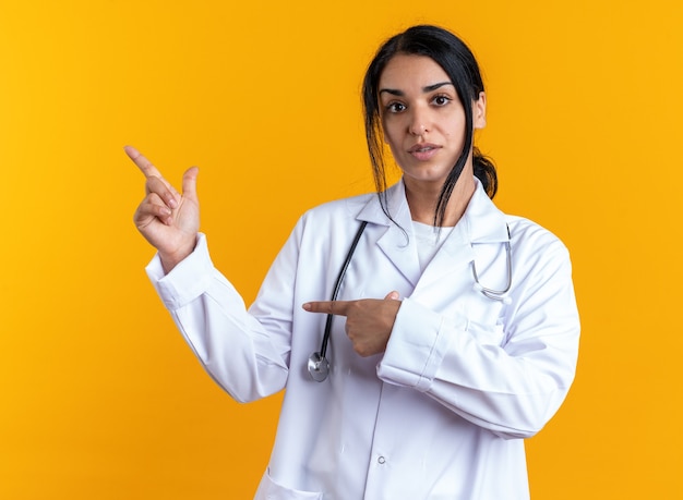 Молодая женщина-врач в медицинском халате со стетоскопом указывает сбоку, изолированную на желтом фоне с копией пространства Бесплатные Фотографии