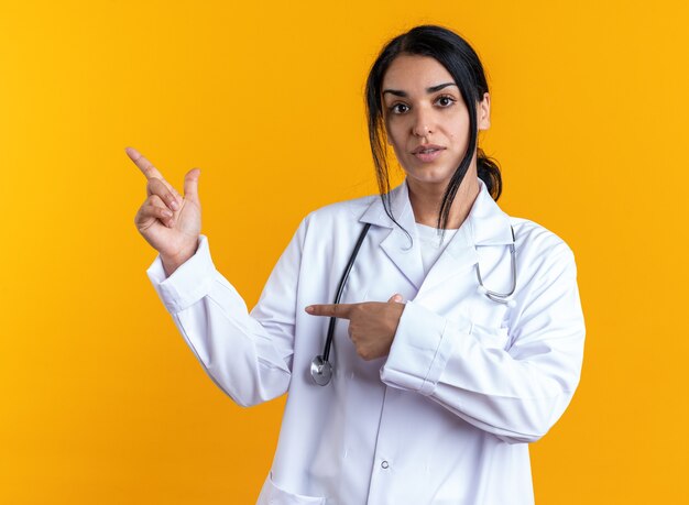 Young female doctor wearing medical robe with stethoscope points at side isolated on yellow background with copy space