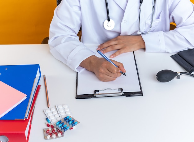 Young female doctor wearing medical robe with stethoscope sits at table with medical tools writing something on clipboard isolated on yellow background