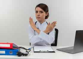 Free photo young female doctor wearing medical robe with stethoscope sitting at desk work on computer with medical tools gesturing no on isolated white wall with copy space