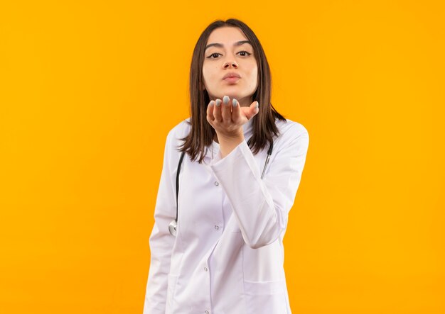 Young female doctor in white coat with stethoscope around her neck blowing a kiss with hand in front of her standing over orange wall