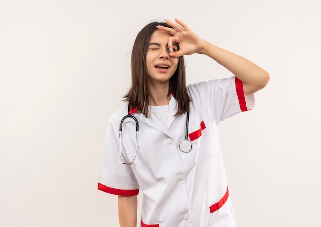 Молодая женщина-врач в белом халате со стетоскопом на шее делает знак ОК, глядя сквозь эту песню, стоя над белой стеной