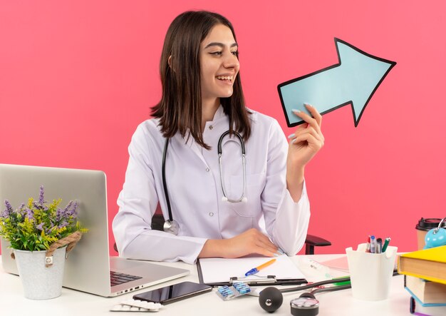 Young female doctor in white coat with stethoscope around her neck holding blue arrow looking aside smiling sitting at the table with laptop over pink wall