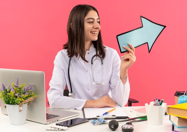 Young female doctor in white coat with stethoscope around her neck holding blue arrow looking aside smiling sitting at the table with laptop over pink wall