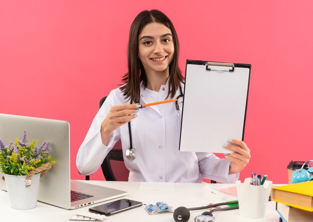분홍색 벽에 노트북과 함께 테이블에 앉아 웃고 그것에 펜으로 가리키는 빈 페이지 클립 보드를 들고 그녀의 목 주위에 청진기와 흰색 코트에 젊은 여성 의사