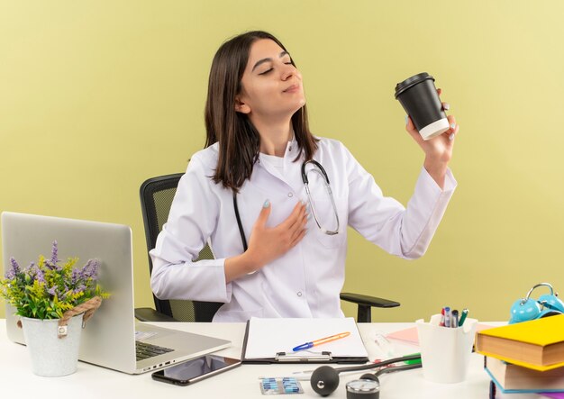 Young female doctor in white coat with stethoscope around her neck holding coffee cup feeling positive emotions sitting at the table with laptop over light wall