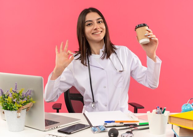 Young female doctor in white coat with stethoscope around her neck holding coffee cup showing ok sign smiling sitting at the table with laptop over pink wall