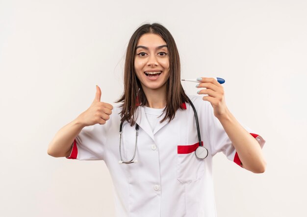 Молодая женщина-врач в белом халате со стетоскопом на шее, держащая цифровой термометр, улыбается, показывает палец вверх, стоя над белой стеной