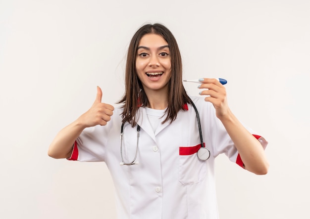Молодая женщина-врач в белом халате со стетоскопом на шее, держащая цифровой термометр, улыбается, показывает палец вверх, стоя над белой стеной