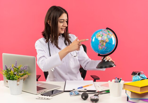 Молодая женщина-врач в белом халате со стетоскопом на шее держит глобус, глядя с улыбкой на лице, сидя за столом с ноутбуком над розовой стеной