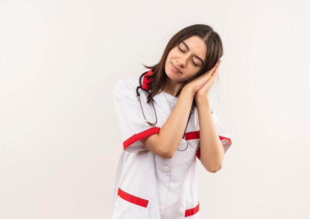 Young female doctor in white coat with stethoscope around her neck holding palms together leaning head on palms with closed eyes wants to sleep looking tired standing over white wall