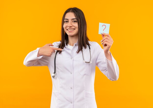 Молодая женщина-врач в белом халате со стетоскопом на шее держит бумагу с напоминанием с вопросительным знаком, указывая пальцем на нее, улыбаясь, глядя вперед, стоя над оранжевой стеной