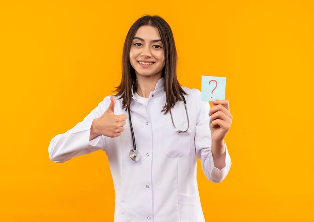 Young female doctor in white coat with stethoscope around her neck holding reminder paper with question mark smiling looking to the front showing thumbs up standing over orange wall