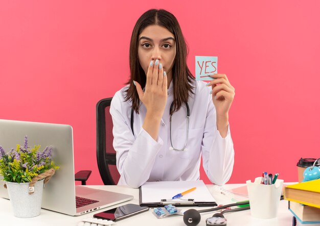 Young female doctor in white coat with stethoscope around her neck holding reminder paper with word yes looking surprised covering mouth with hand sitting at the table with laptop over pink wall