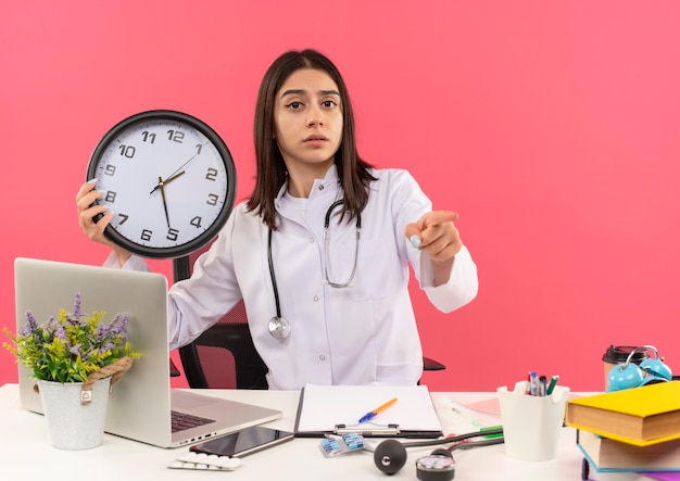 Young female doctor in white coat with stethoscope around her neck holding wall clock pointing with finger to the front looking confused sitting at the table with laptop over pink wall