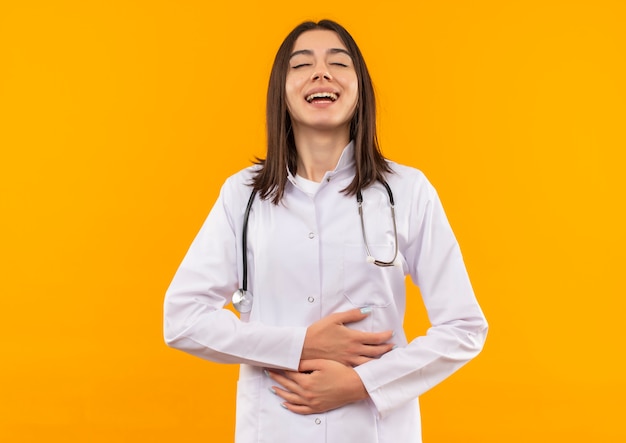 Young female doctor in white coat with stethoscope around her neck laughing out touching her belly standing over orange wall