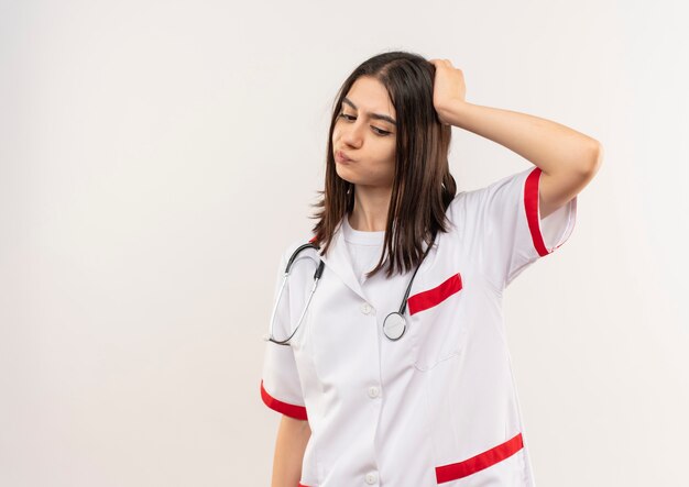 Молодая женщина-врач в белом халате со стетоскопом на шее, озадаченно глядя в сторону, стоя у белой стены