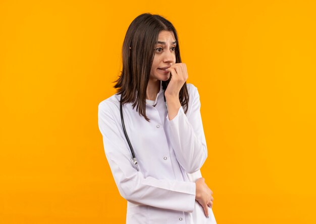 Молодая женщина-врач в белом халате со стетоскопом на шее смотрит в сторону напряженно и нервно, стоя у оранжевой стены