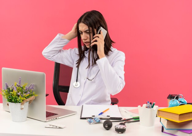 ピンクの壁の上のラップトップでテーブルに座って携帯電話で話している間彼女の首の周りに聴診器を持った白衣の若い女性医師