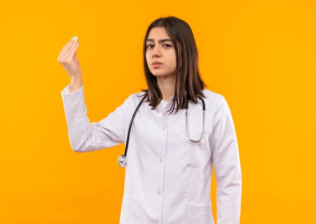 Young female doctor in white coat with stethoscope around her neck looking to the front displeased gesturing with hand standing over orange wall