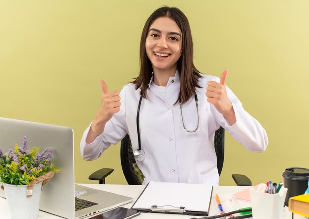Young female doctor in white coat with stethoscope around her neck looking to the front smiling cheerfully showing thumbs up, sitting at the table with laptop over light wall
