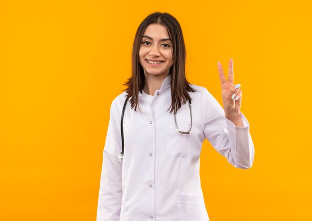 Young female doctor in white coat with stethoscope around her neck looking to the front smiling cheerfully showing victory sign standing over orange wall