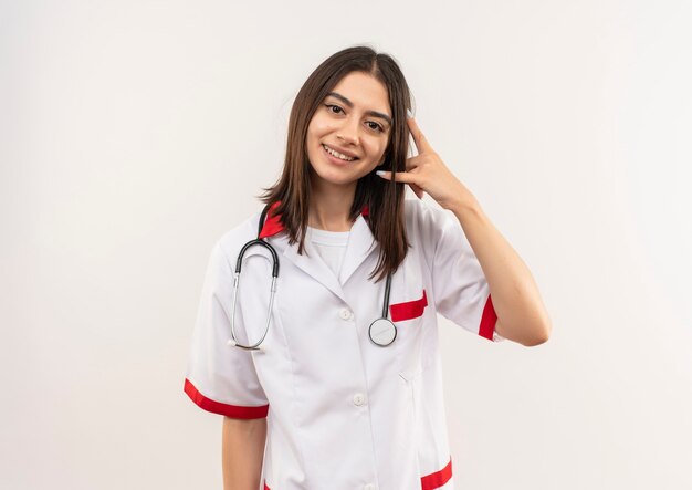 首に聴診器を持った白いコートを着た若い女性医師が正面を向いて笑って、白い壁の上に立っているジェスチャーを呼んでください