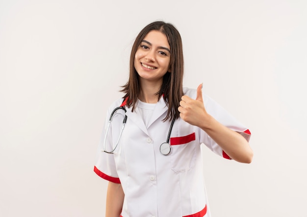 Молодая женщина-врач в белом халате со стетоскопом на шее, глядя вперед, улыбаясь, показывает палец вверх, стоя над белой стеной