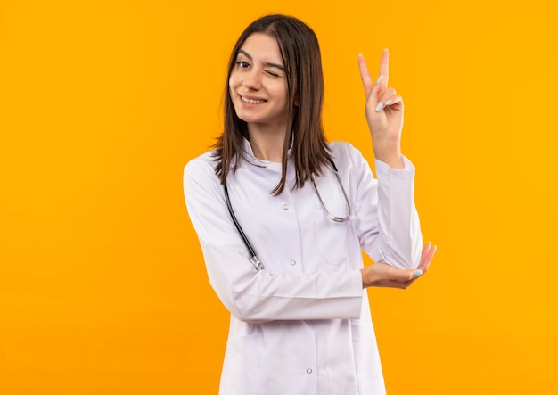 首の周りに聴診器を持った白いコートを着た若い女性医師が笑顔でウインクしてオレンジ色の壁の上に立っている勝利のサインを示しています