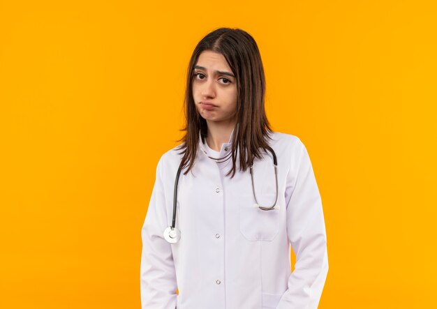 오렌지 벽 위에 서있는 얼굴을 찡그린 얼굴로 앞으로 찾고 그녀의 목에 청진기와 흰색 코트에 젊은 여성 의사