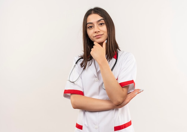 Young female doctor in white coat with stethoscope around her neck looking to the front with hand on chin looking confident thinking standing over white wall